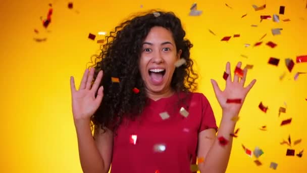 Young latin girl with curly hair dancing and having fun in confetti rain on yellow background. Woman celebrating, depicts joy and happiness. Success, victory, holiday concept. - Filmagem, Vídeo