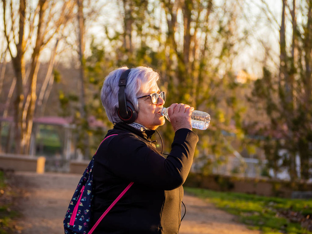 Una mujer mayor con cabello blanco escuchando música con auriculares y bebiendo agua de una botella de plástico
 - Foto, imagen