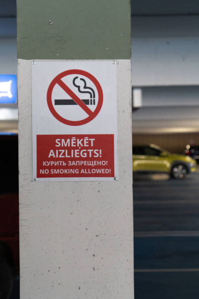Vermelho Proibido fumar sinal permitido em três idiomas em um estacionamento subterrâneo com carros visíveis no fundo
 - Foto, Imagem