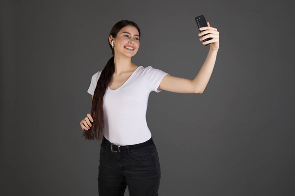 attraktives brünettes Mädchen in weißem T-Shirt isoliert über grauem Hintergrund, das Handy lächelnd anschaut - Foto, Bild