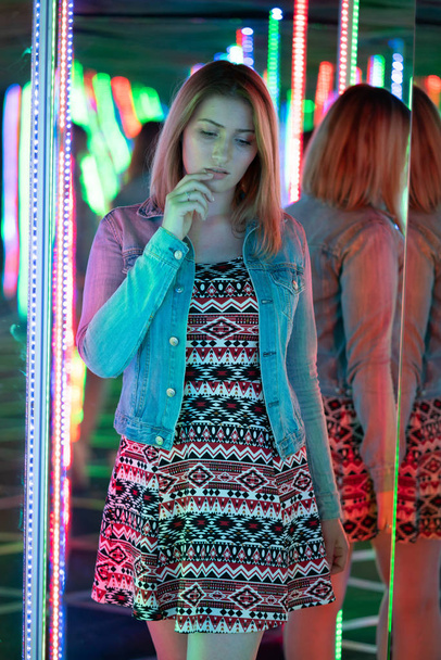 sweet caucasian girl walks in a mirror maze with colorful diodes and enjoys an unusual attraction room in the city - Photo, Image