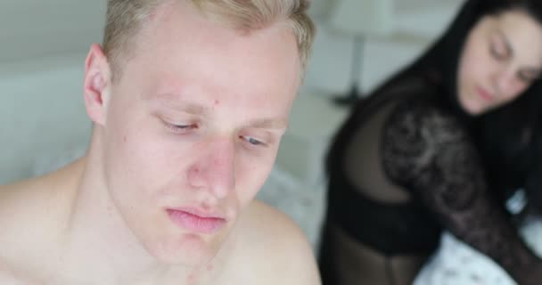 Handsome blond blue eyed young man feels depressed during bedroom crisis, woman in black underwear sitting in background - Footage, Video