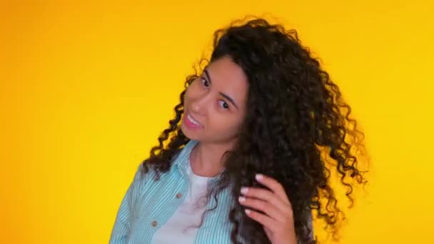 Portrait of young beautiful girl corrects her curls on yellow background. Trendy cute woman smiling to camera. Studio footage, latin or hispanic positive female - Кадры, видео