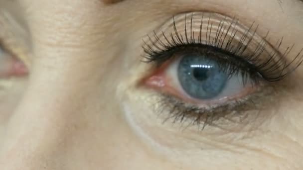 Profesjonalny makijaż artysta laski fałszywe rzęsy na niebieskich oczach dorosłego w średnim wieku kobieta bliska widok - Materiał filmowy, wideo