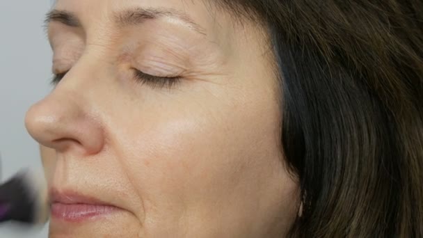 Make up καλλιτέχνη κάνει μακιγιάζ με το κονσίλερ επισήμανσης σε μεσήλικες γυναίκα με μπλε μάτια. Μακιγιάζ ηλικίας. Κοντινό πορτρέτο. Εφαρμόστε σκόνη με βούρτσα - Πλάνα, βίντεο