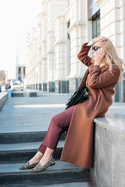 Stylish fashionable blonde woman wearing coat and sunglasses speaking on mobile phone, street style photo - Foto, Imagen