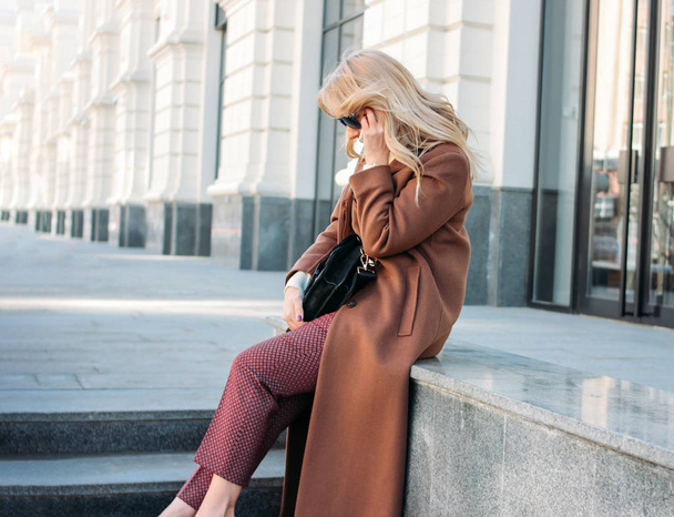 Stylové módní blondýna žena nosí kabát a brýle mluvit na mobilním telefonu, pouliční styl photo - Fotografie, Obrázek