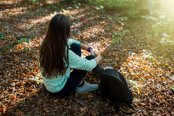 Young woman resting, sitting after a walk in the forest, with backpack on sunset light in the autumn season. - Фото, зображення
