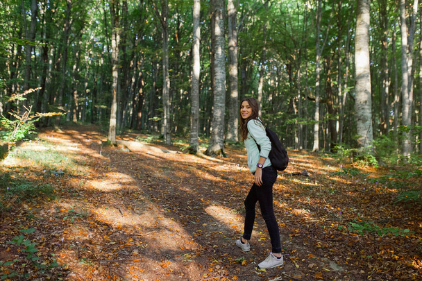 Młoda kobieta biorąca spacer w lesie, przewożąca plecak w lesie na świetle słońca w sezonie jesiennym. - Zdjęcie, obraz