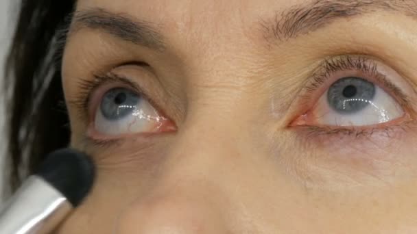 Make up καλλιτέχνη κάνει μακιγιάζ με το κονσίλερ επισήμανσης σε μεσήλικες γυναίκα με μπλε μάτια. Μακιγιάζ ηλικίας. Κοντινό πορτρέτο. Εφαρμόστε σκόνη με βούρτσα - Πλάνα, βίντεο