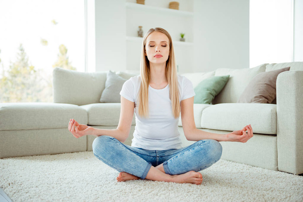 Портрет серьезной дамы Millennial тренировки йоги упражнения медитативный поиск спокойствие сидеть на полу лотос представить воображение красивый диван одет джинса современный модный наряд в гостиной
 - Фото, изображение