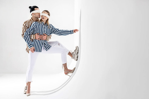 Couple wearing white trousers posing near white wall in blindfolds - 写真・画像