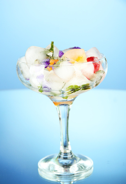 Ice cubes with flowers and herbs and fruits inside in cocktail glass, on blue background - Photo, image