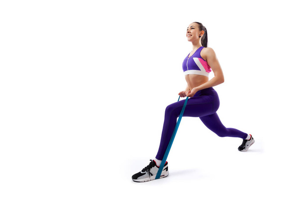Uma treinadora de cabelos escuros em um top curto roxo esportivo e leggings ginásio faz lunges com elásticos esporte fitness em um fundo isolado branco em estúdio
  - Foto, Imagem
