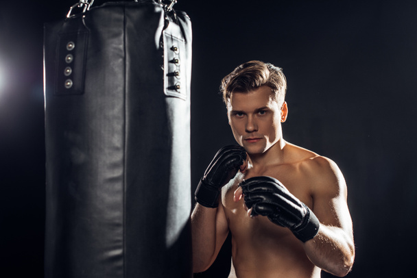 Strong boxer in gloves standing near punching bag and looking at camera - Фото, зображення