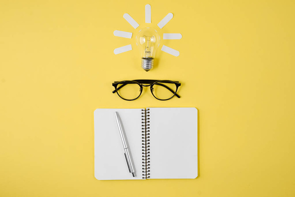 Flat lay, top view, copy space. Financial planning table top with pen, notepad, eyeglasses and light bulb on yellow background. Concept brainstorming and new idea - Фото, зображення