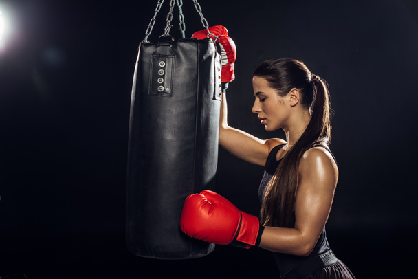 Vista lateral del boxeador femenino en guantes de boxeo rojos entrenando con saco de boxeo
 - Foto, Imagen