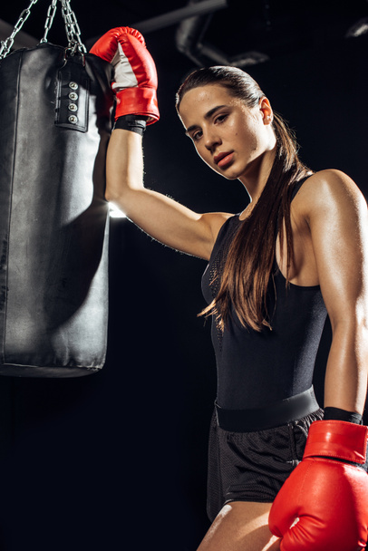 Женщина-боксер в красных боксерских перчатках стоит рядом с боксерской грушей
 - Фото, изображение