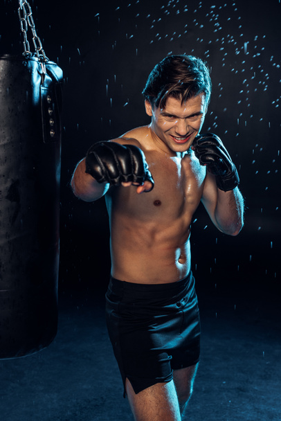 Hymyilevä nyrkkeilijä käsineissä seisoo lähellä nyrkkeilysäkkiä vesipisaroiden alla mustalla
 - Valokuva, kuva