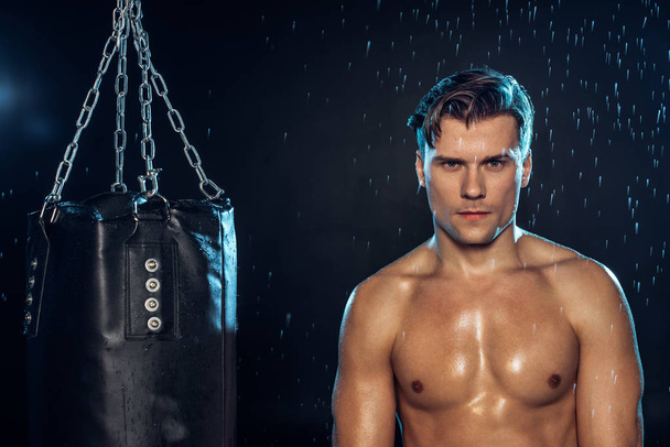 Front view of boxer standing near punching bag under water drops on black - Fotografie, Obrázek