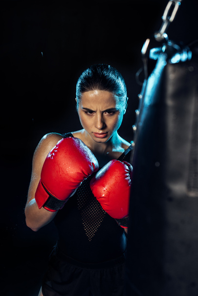 Boxeador concentrado en guantes de boxeo rojos mirando el saco de boxeo en negro
 - Foto, Imagen