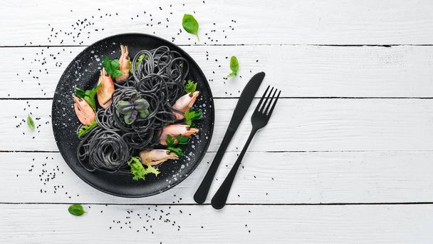 Black pasta with shrimps on a black plate. Top view. On a white wooden background. Free copy space. - Photo, image