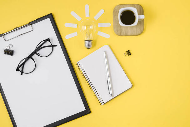 Creative flat lay concept top view of workspace desk styled design office supplies with pen, notepad, eyeglasses, cup coffee and light bulb on a yellow background. Concept brainstorming - Foto, imagen