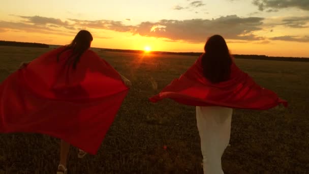 Moeder en dochter spelen super helden in rode mantels bij zonsondergang in het Park. Meisjesspelen super helden lopen over het veld tegen de blauwe hemel. concept van een gelukkige familie. concept Woman, redder, winnaar - Video