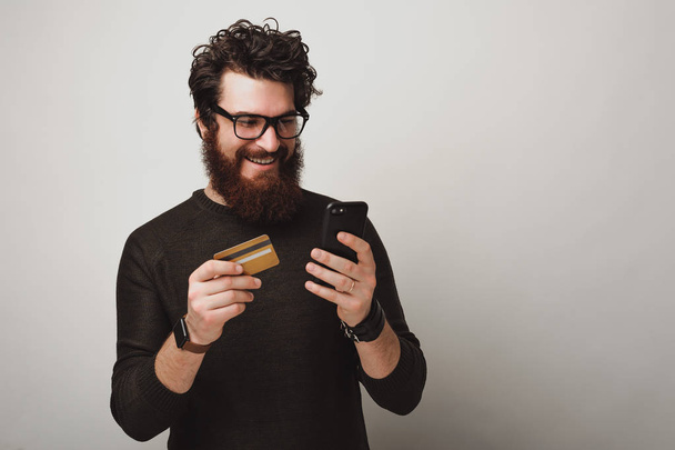 Όμορφος διαφήμιση χαρούμενο μούσι άνθρωπος χρησιμοποιώντας το τηλέφωνο και τη κατοχή πιστωτική κάρτα για την ηλεκτρονική πληρωμή - Φωτογραφία, εικόνα