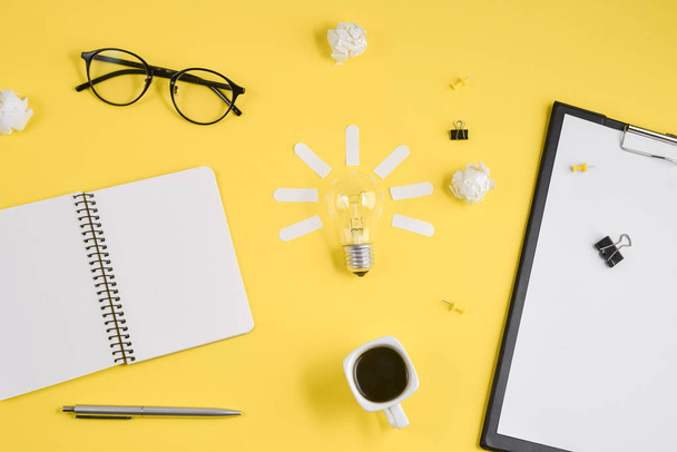 Office workspace with blank clip board, office supplies, pen, notepad, eyeglasses, coffee cup and light bulb on yellow background. Concept brainstorming and new idea - Foto, imagen