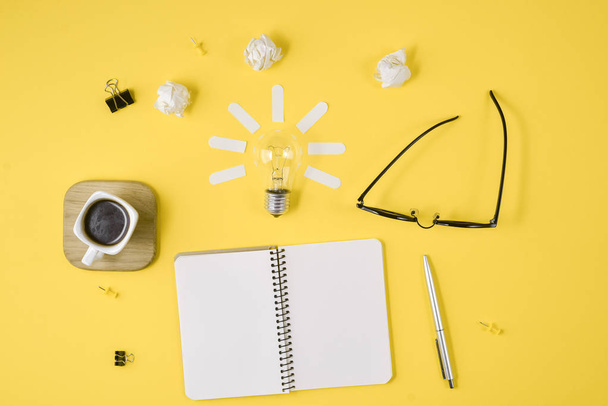 Flat lay financial planning brainstorming messy table top image with blank clip board, office supplies, pen, notepad, eyeglasses, cup of coffee, light bulb on yellow background. - Foto, imagen
