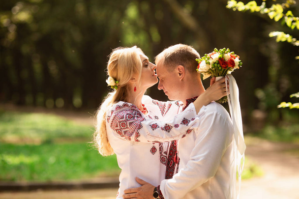 A young couple in a traditional Ukrainian clothing whith bouquet walking in the sunny park - Photo, Image