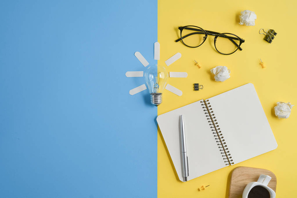 Financial planning table top with blank clip board, office supplies, pen, notepad, eyeglasses, coffee cup, light bulb on yellow and blue background. Conceptual brainstorming still life - Foto, imagen