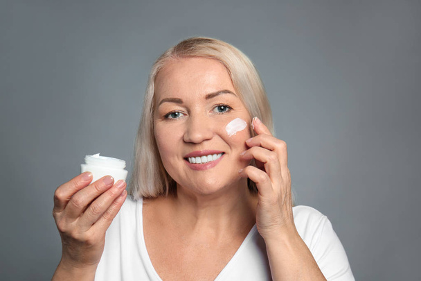 Mature woman applying facial cream on grey background - Photo, Image