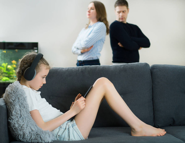 Little girl in the headphones using tablet pc while her parents in quarrel in the background. - Photo, Image