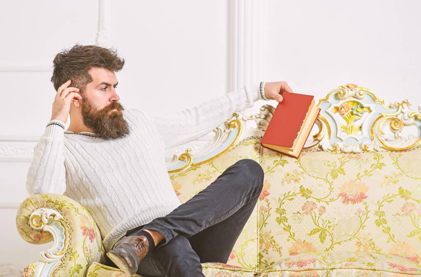 Man with beard and mustache spends leisure with book. Scientist, professor on strict face analyzing literature. Self education concept. Lecturer sit on sofa and holds book, white wall background - Photo, image
