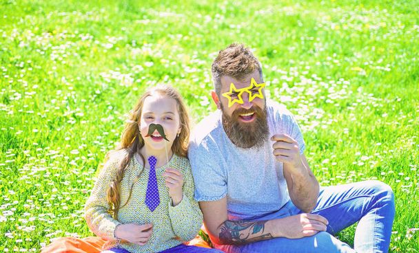 Best friends concept. Dad and daughter sits on grass at grassplot, green background. Family spend leisure outdoors. Child and father posing with eyeglases, mustache and tie photo booth attributes - Fotografie, Obrázek