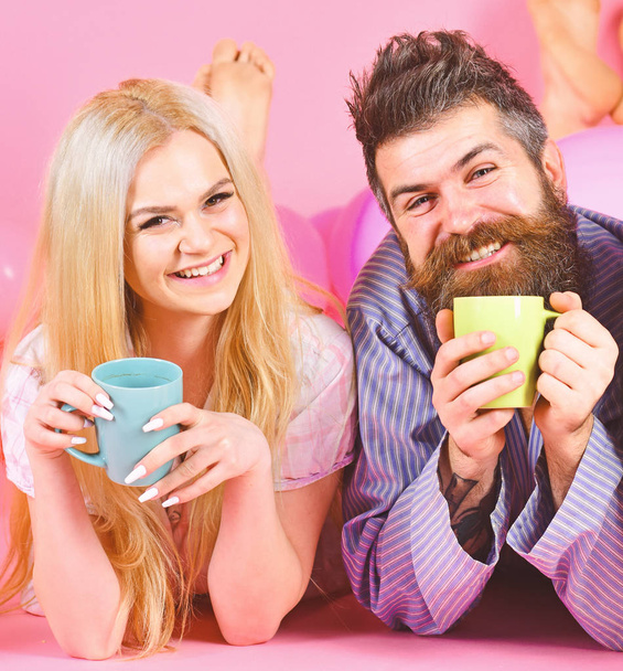 Couple relax in morning with coffee. Family tradition concept. Couple in love drink coffee in bed. Man and woman in domestic clothes, pajamas. Man and woman on smiling faces lay, pink background - Photo, image