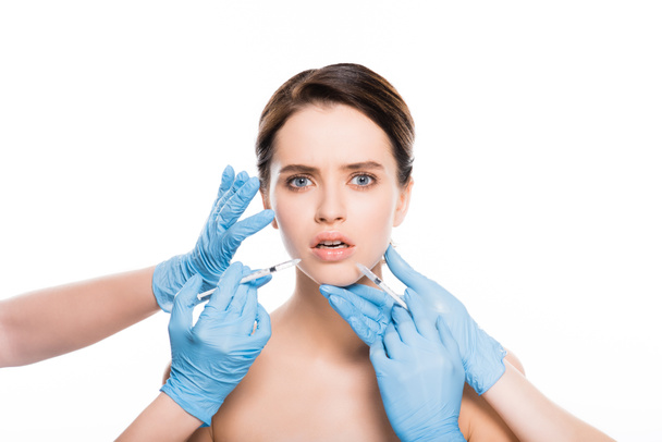 oříznutý pohled kosmetologů v latexových rukavicích dotyků šokované dívky při držení injekční stříkačky izolované na bílém - Fotografie, Obrázek