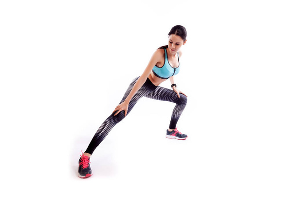 Une femme aux cheveux bruns entraineuse dans un haut court sportif et leggings de gymnastique fait des fentes par le côté des pieds, les mains sont tendues sur le côté sur un fond blanc isolé en studio
  - Photo, image