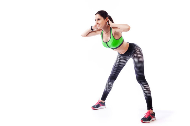 A dark-haired woman coach in a sporty short top and gym leggings makes bends forward, arms behind head, legs wide apart  on a  white isolated background in studio  - Foto, imagen