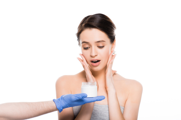 abgeschnittene Ansicht des plastischen Chirurgen in Latex-Handschuh-Aufbewahrungsbehälter mit kosmetischer Creme in der Nähe überrascht Mädchen isoliert auf weiß  - Foto, Bild