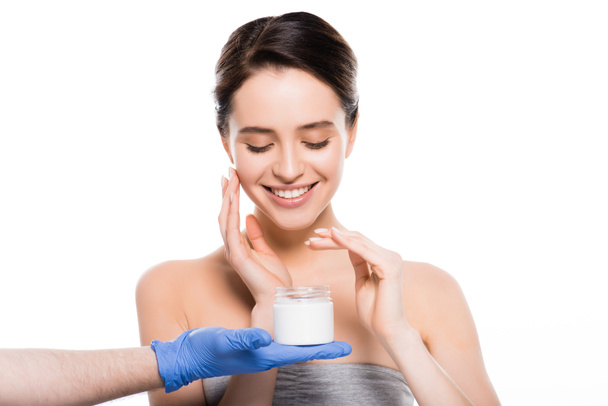 περικομμένη όψη του πλαστικού χειρουργού σε γάντια λατέξ κρατώντας δοχείο με καλλυντική κρέμα κοντά ευτυχισμένο κορίτσι απομονωθεί σε λευκό  - Φωτογραφία, εικόνα