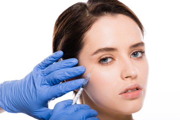 περικομμένη όψη του αισθητολόγου σε μπλε λατέξ γάντια καουτσούκ κρατώντας σύριγγα κοντά στο πρόσωπο της νεαρής γυναίκας που απομονώνεται σε λευκό  - Φωτογραφία, εικόνα