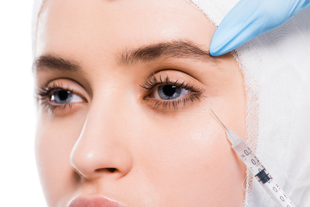 abgeschnittene Ansicht des Kosmetikers mit Latexhandschuhen, die Spritze in der Nähe des Auges der Frau mit bandagiertem Kopf, isoliert auf weiß  - Foto, Bild