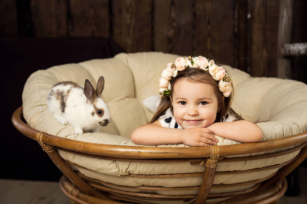 Portrait of a beautiful little girl in a wreath of flowers. A girl with a cute fluffy white Easter bunny - Фото, зображення