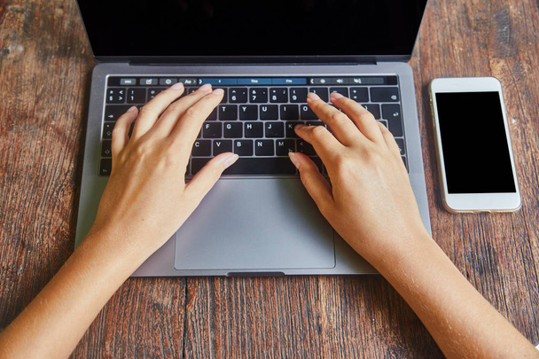 Visão superior da bela mulher mãos digitando no teclado do laptop colocado no desktop de escritório de madeira com smartphone em branco
. - Foto, Imagem