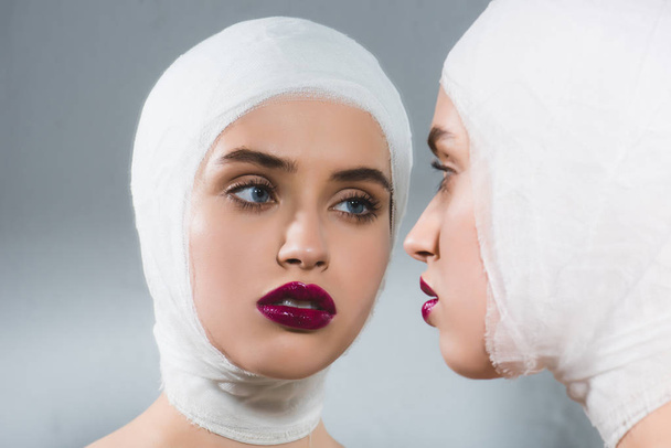 junge Frau mit bandagiertem Kopf in der Nähe von Spiegel auf grau  - Foto, Bild