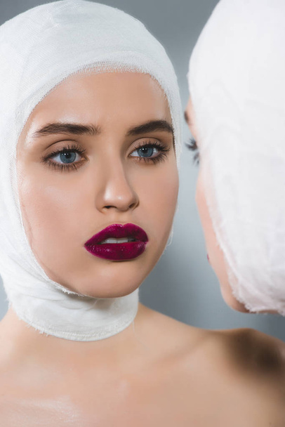 young pretty woman with bandaged head near mirror on grey  - Фото, изображение