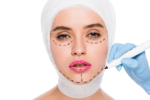 vista recortada de cirujano plástico en guante de látex con rotulador cerca de chica atractiva con marcas en la cara aislada en blanco
  - Foto, imagen